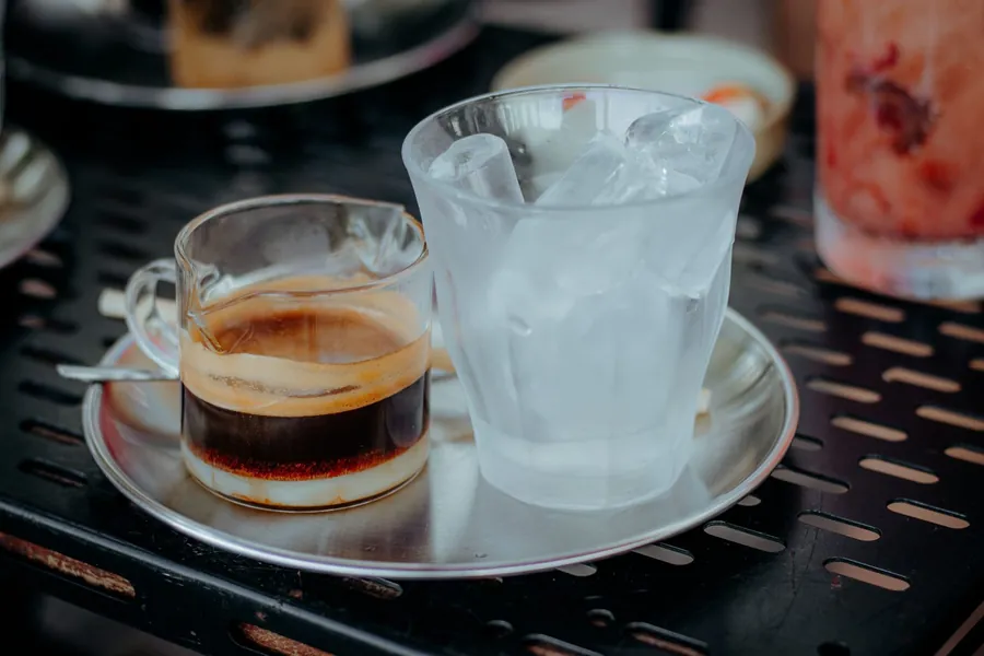 A shot of espresso beside a glass of ice on a steel tray